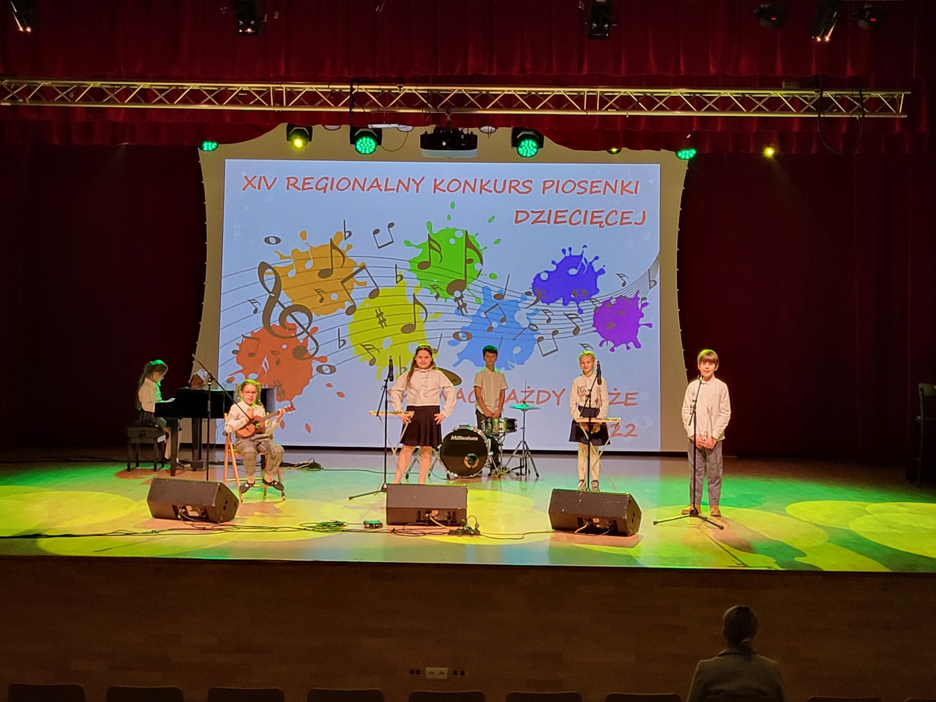 Sześcioro uczniów tworzących zespół muzyczny stoi na scenie. Uczniowie stoją przy mikrofonach i instrumentach. W tle widać widoczny napis "XIV Regionalny Konkurs Piosenki Dziecięcej".