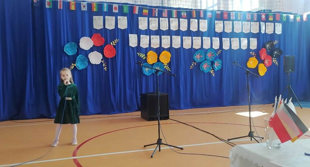 Na zdjęciu widać dziewczynkę w zielonej sukience, śpiewającą do mikrofonu, obok dwa stojaki na mikrofony i głośnik. W tle niebieskie płótno ozdobione dużymi kolorowymi kwiatami i flagami różnych krajów.