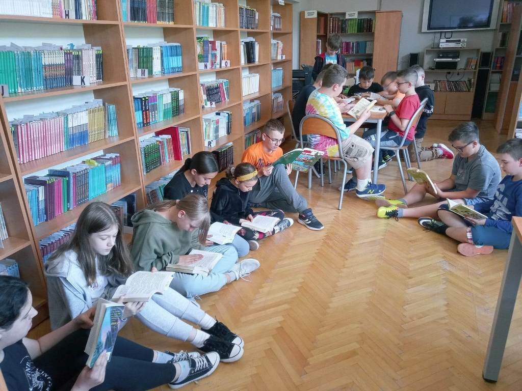 Na zdjęciu widzimy dużą grupę uczniów klasy czwartej w bibliotece szkolnej. Część z nich siedzi na podłodze, a inni przy stoliku. Wszyscy trzymają w rękach otwarte książki. Po lewej stronie znajdują się regały biblioteczne z kolorowymi krzesłami.
