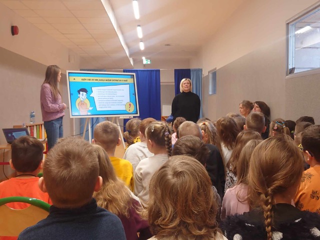 grupa dzieci siedzi wpatrując się w prezentację wyświetlaną na tablicy interaktywnej, przy której stoją dwie panie nauczycielki