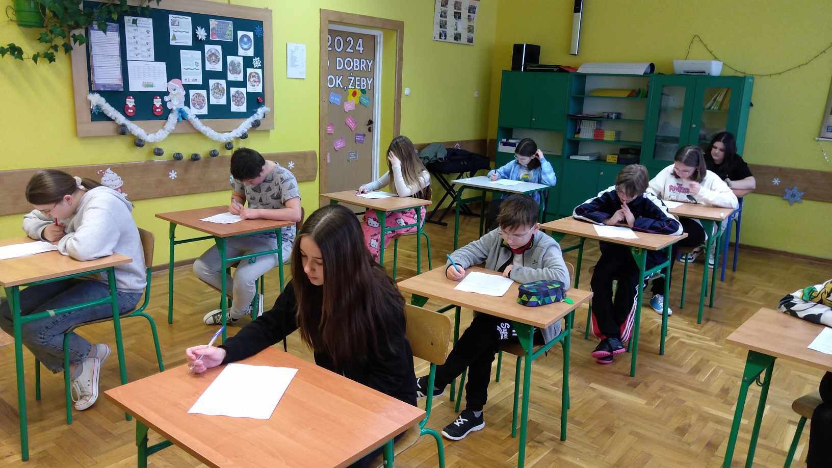 W sali lekcyjnej siedzą uczniowie przy stolikach rozwiązując test wiedzy.