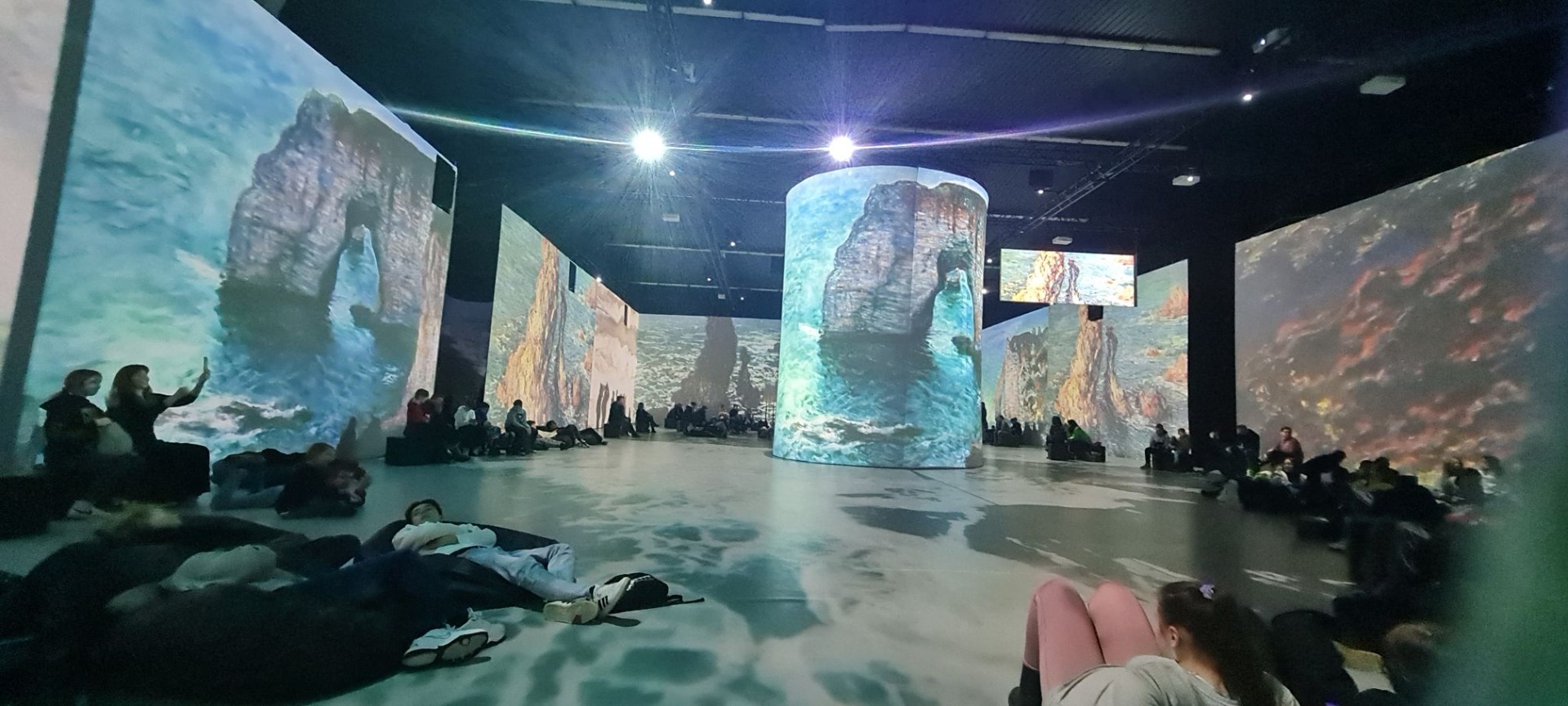 Obraz przedstawiający morze z formacjami skalnymi wyświetlony na wielkoformatowych powierzchniach. Widoczne sylwetki osób oglądających wystawę.