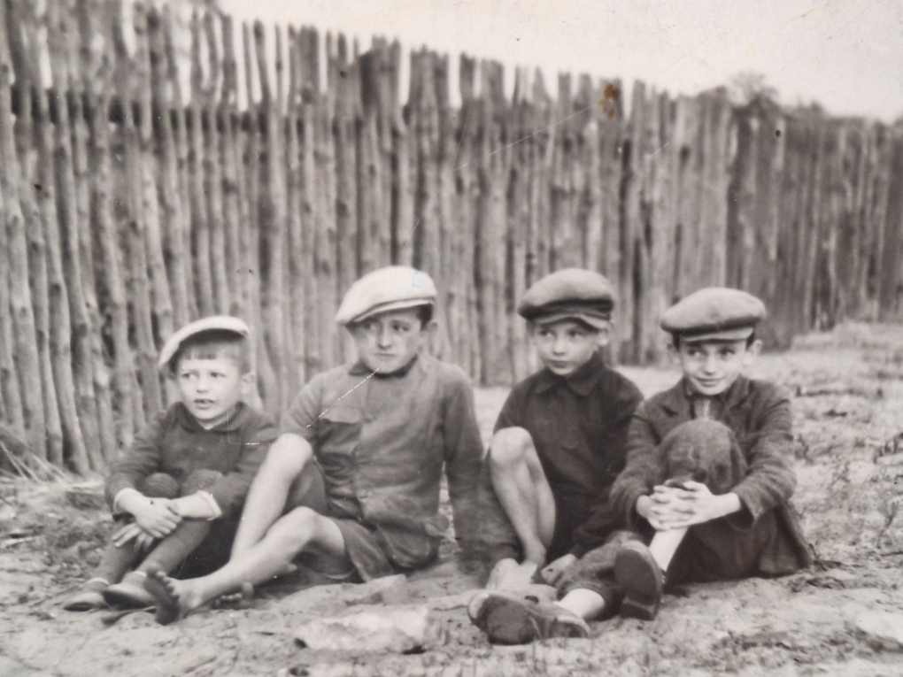 Czarno białe przedwojenne zdjęcie pokazujące 4 ok 8 letenich chłopców w dawnych strojach. Siedzą przed płotem zbitych z desek.