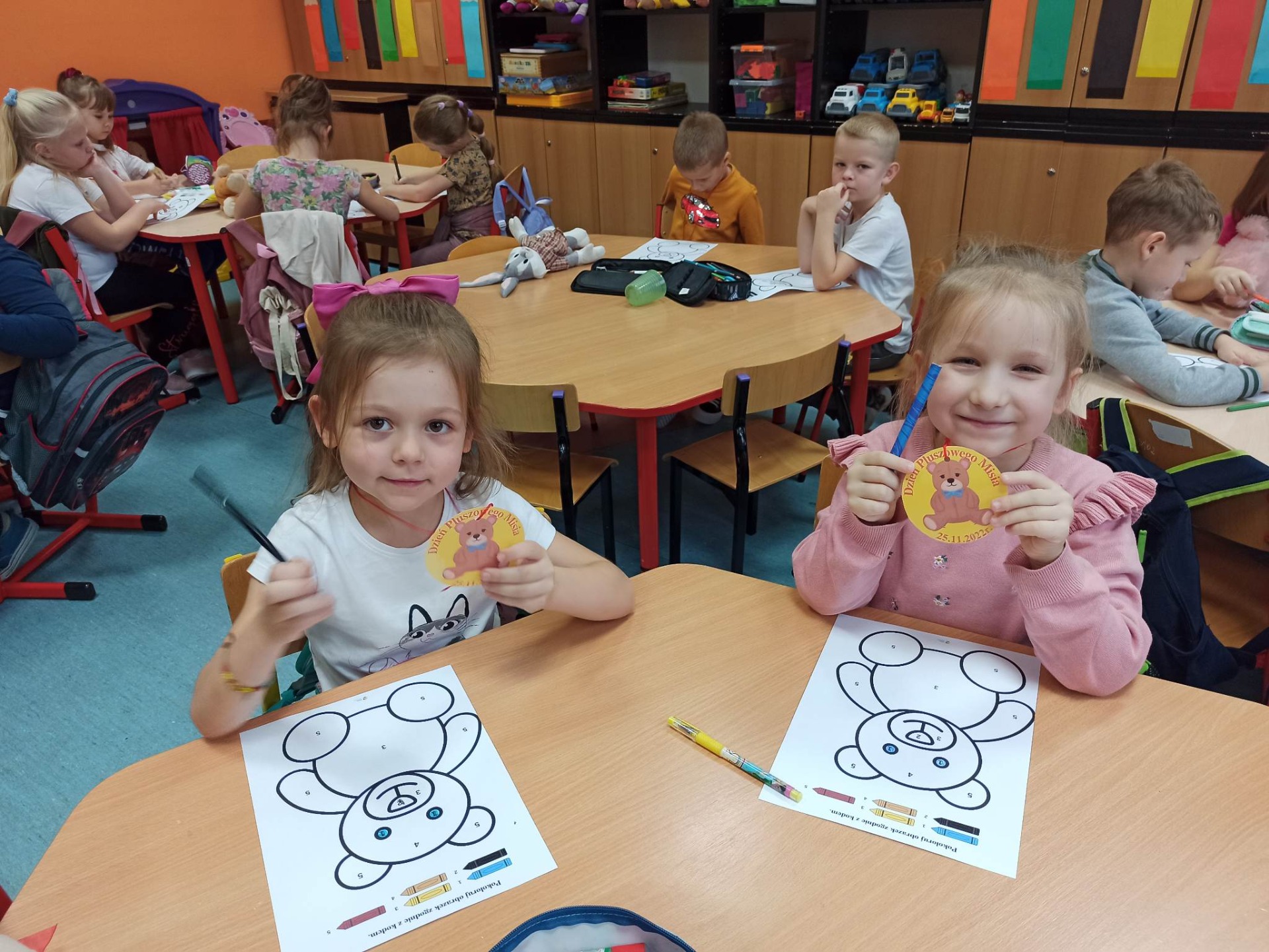 Dzieci siedzą przy stolikach w sali przedszkolnej i kolorują rysunki z misiem