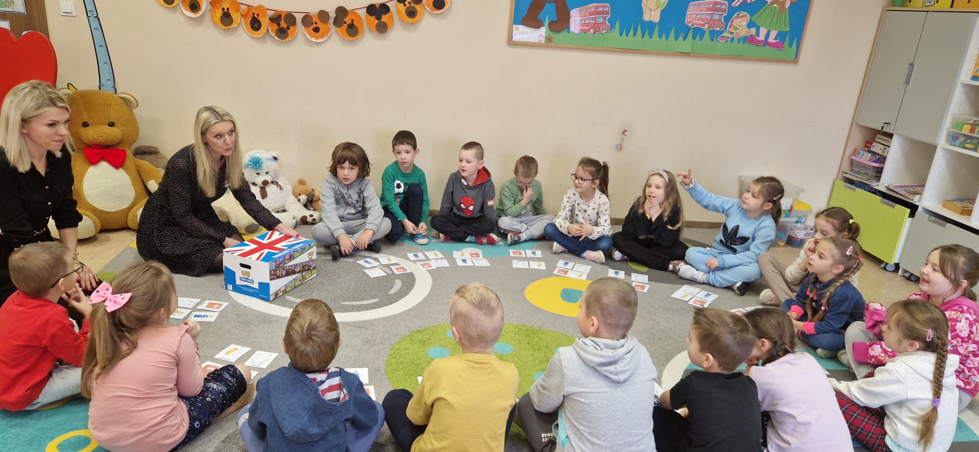 Zdjęcie przedstawia dzieci z oddziału przedszkolnego A, które siedzą na dywanie w okręgu wraz z wychowawczynią i panią przeprowadzającą warsztaty językowe.