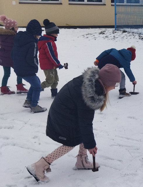 Na zdjęciu znajdują się dzieci z oddziału przedszkolnego.  Dzieci trzymają drewniane stemple, które odbijaja na śniegu. Dzieci są uśmiechnięte. Zdjęcie zostało zrobione na placu szkolnym.