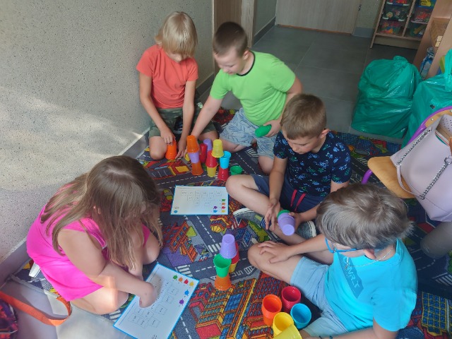 Uczniowie siedzą na dywanie. Nachyleni nad kartą pracy, rozwiązują ją układając kolorowe kubeczki.