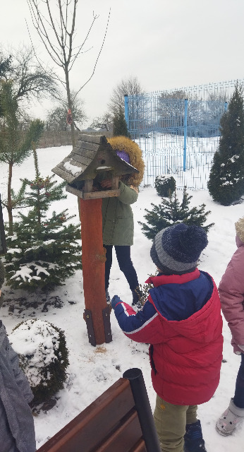 Na zdjęciu znajdują się dzieci z oddziału przedszkolnego.  Dzieci trzymają drewniane stemple, które odbijaja na śniegu. Dzieci są uśmiechnięte. Zdjęcie zostało zrobione na placu szkolnym.
